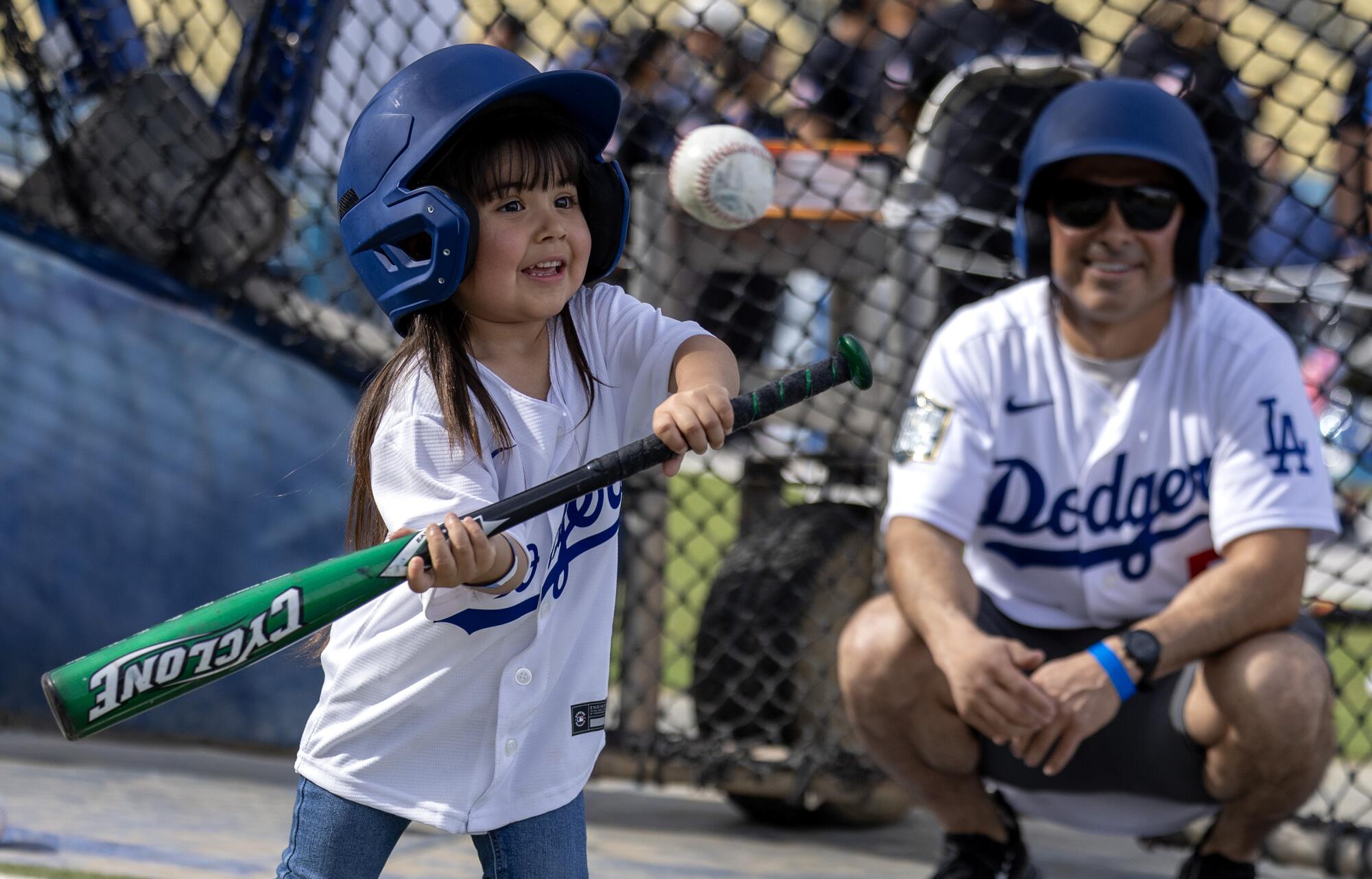 A little girl in a Dodgers shirt holds a baseball bat, ready to swing at an incoming ball as her father watches. 